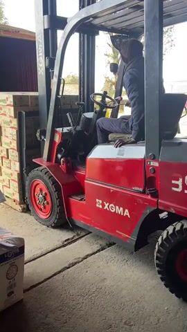 A person wearing a blue jumpsuit and a black hood is operating a red XGMA forklift. The forklift is positioned to lift a pallet of cardboard boxes, which are stacked high and appear to be inside a shipping container. The forklift's forks are engaged with the pallet. In the foreground, a white bag with green lettering is visible on the ground. The scene appears to be an outdoor industrial or storage area with concrete flooring and some vegetation in the background. The forklift's mast is raised, indicating the process of lifting or moving the pallet. The driver is looking forward, presumably guiding the forklift's movement.