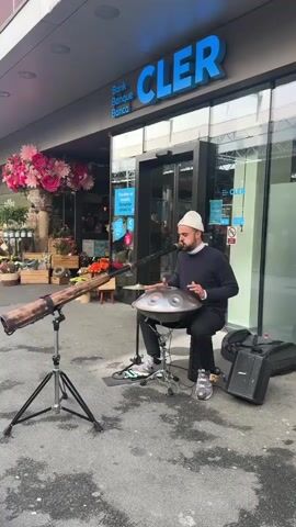 A man is busking in front of a Cler bank branch. He sits playing a handpan, and is accompanied by a didgeridoo and a foot pedal. The didgeridoo is mounted on a tripod. There are flowers in front of the bank branch, and a passerby is visible. The man wears a navy sweater, black pants, a white beanie, and colorful sneakers. He has a dark beard. A microphone is positioned in front of him, and a portable speaker and bag are nearby.