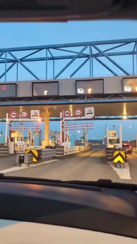 The video appears to be recorded from inside a vehicle, looking forward through the windshield. The vehicle is approaching a toll booth, indicated by the overhead signage, barriers, and payment stations. The sky is a deep blue, suggesting it is either dawn or dusk. Several toll lanes are visible, some with red lights indicating they are closed, and others with green lights, likely open. The price of 6.10€ is displayed on a sign at one of the lanes. The camera pans slightly to the right, showing the interior of the car, including the dashboard and steering wheel. The vehicle continues to move forward into one of the toll lanes, and the video ends as it appears to be passing through.
