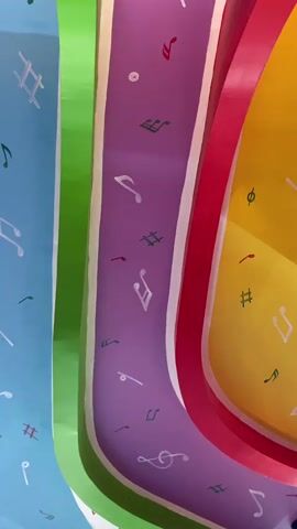 The video pans across a ceiling decorated with colorful, wavy stripes of blue, green, purple, pink, and yellow. Each stripe is adorned with various musical notes and symbols, such as treble clefs, eighth notes, and hash symbols, painted in white and red. Below the ceiling, a textured, dark brown material, possibly woven reeds or branches, is visible. Further down, a large window with red framing reveals a lush green outdoor environment with trees and foliage. A glimpse of a motorcycle is visible outside through the window. The interior also shows parts of what appears to be a cafe or dining area, with a dark chair and a table visible near the window. The lighting in the scene suggests it is daytime.