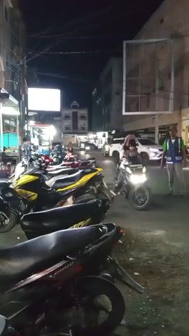 The video captures a nighttime scene on a street in Bandarbireuen, Indonesia. The setting appears to be a commercial area with several buildings lining the road, some of which are illuminated. Parked motorcycles, a common mode of transport in the region, are prominently featured in the foreground and along the side of the street. The lighting suggests it is evening or nighttime, with artificial lights from buildings and streetlights illuminating the area.

The activity centers around a person in a high-visibility vest, possibly a traffic controller or security personnel, walking across the street. Another person is seen on a scooter, preparing to move. In the background, other vehicles, including cars and more scooters, are visible, suggesting ongoing traffic. The overall atmosphere appears to be that of a busy street at night, with people going about their activities.

Notable details include the presence of various types of motorcycles, some with bright yellow accents, and the layered clothing of the individuals, which might indicate cooler evening temperatures or a cultural norm. The buildings show a mix of architectural styles, with some appearing older and others more modern. A large, somewhat weathered billboard structure is visible on one of the buildings. The street surface is asphalt, with some debris or unevenness. The weather appears to be clear, given the visible sky.

There is no easily discernible text in the video, though some signs on buildings may be present but are too blurry or distant to read. The scene conveys a sense of everyday life in an Indonesian town at night, with a focus on street activity and transportation.