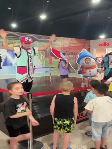 A group of children and adults are gathered around a raised platform in what appears to be a shopping mall. On the platform, a woman in a pink hat and a turquoise vest is interacting with two children holding a cutout of a snowman. The snowman cutout has a hole for a face. Another person in a bright turquoise and black costume with a TikTok logo is dancing on the platform. Several children are watching from the front, some are looking towards the platform, and others are looking at each other. In the background, there is a colorful mural of a playground and a store called Zolla. The overall atmosphere seems to be an organized event or performance for children.