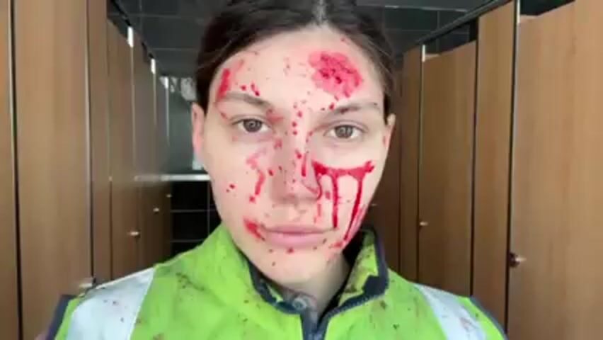 The image shows a woman in a green safety vest standing in what appears to be a public restroom with multiple stalls. She is covered in what seems to be blood splatters and smears on her face and vest. The blood is concentrated around her forehead and dripping down her face. She is looking directly at the camera with a serious expression. The background is a neutral-toned restroom with wooden stall doors.