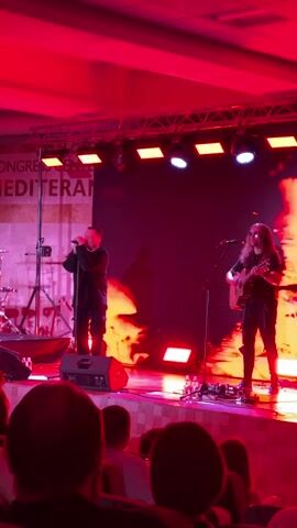 The video shows a live performance on a stage. The stage is lit with red and pink lights. In the center of the stage, a male singer wearing a black hoodie and black pants stands behind a microphone. To the right of the singer, a male guitarist with long hair, wearing a black shirt and black pants, plays an electric guitar. Behind them, a large screen displays a fiery, abstract image. To the left of the stage, drums and other musical equipment are visible. In the foreground, the backs of the heads of audience members are visible. A banner with text that includes "CONGRESO MEDITERAN" is visible on the left side of the stage.