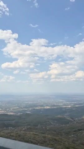 The video shows a panoramic view of a valley from a high vantage point. The sky is blue with scattered white clouds. The landscape below is a mix of green hills and fields, with buildings and roads visible in the distance. The foreground is dominated by a metallic barrier, possibly a guardrail, suggesting the video was taken from a road or viewpoint. The overall impression is one of a vast, open landscape under a clear sky.