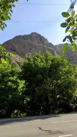 The video pans from left to right, showing a mountain in the background and trees in the foreground. A road runs across the bottom of the frame. There are power lines visible in the upper right corner of the frame. The sky is blue and clear. The scene appears to be a rural or natural setting.