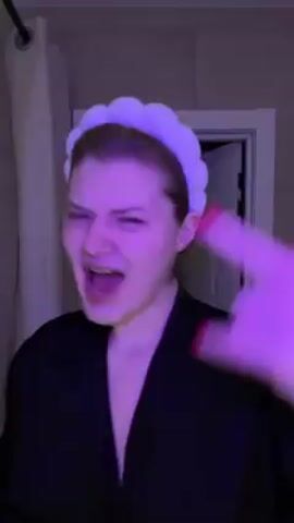 A woman is videoed from the waist up. She wears a white headband and a black robe. She stands in what looks like a bathroom, illuminated by purple light. She makes exaggerated hand gestures while mouthing to a song. The backdrop includes a shower curtain and a partially open door.