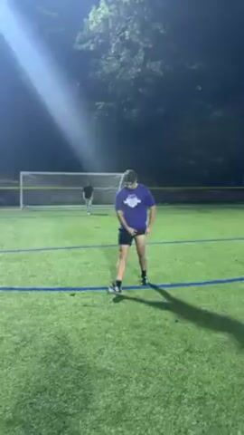 The video shows a man standing on a soccer field with artificial turf under a bright light at night. He is wearing a purple t-shirt with a logo, black shorts, and black socks. There is a soccer goal behind him. The camera pans to the left, showing more of the field.
