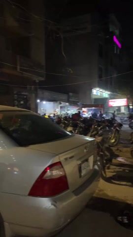 The video shows a street scene at night.  A silver car is parked on the side of the road. There are motorbikes parked near the car. A food shop with a green sign is open and people are standing inside, presumably customers.  A man wearing a black shirt and pants is standing near tables and chairs that appear to be set for outside dining. Other shops can be seen in the background. Streetlights illuminate the area.