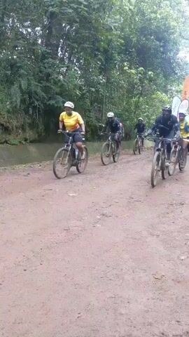 The video shows a group of people participating in a mountain bike race or event on a dirt road surrounded by lush green trees.  Several cyclists are riding their bikes, wearing helmets and athletic gear. They are followed by people on foot wearing vests, some carrying flags or banners with logos and text.  The environment is wooded, giving the impression of a trail in a forest or park.  The road is unpaved and appears to be a natural surface. There are a few spectators. The video seems to be shot from the side of the trail as the event passes.
