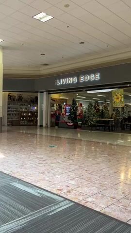 The video pans across a mall concourse. The floor is tiled in a light pink and white pattern. To the right, the store "LIVING EDGE" is visible with Christmas decorations, including trees, in the window. Further down the concourse, a "KIDS ZONE" with a yellow and red inflatable structure is visible. Several people are walking through the mall. A couch area with seating is in the foreground. The lighting suggests daytime.