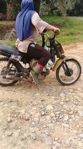 A person wearing a blue headscarf, a light brown shirt, dark pants, pink socks, and green boots is sitting on a yellow motorcycle on a dirt road. The motorcycle is positioned in the center of the frame, with its front wheel slightly turned to the left. The person is holding onto the handlebars, and their body is angled away from the camera, facing towards the right. Behind the motorcycle, there are palm trees and green foliage, indicating an outdoor, possibly rural, environment. The ground is covered with dirt and small stones. The video captures the person sitting still on the motorcycle.
