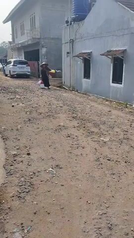 A person wearing a conical hat and dark clothing walks down a gravel road, carrying two white plastic bags and a yellow container in their hands. They are walking towards the right side of the frame. To the left of the road, a white car is parked, and behind it, a two-story building is visible. To the right of the road, a single-story house with gray walls and two windows with awnings is present. The sky is clear and the lighting suggests it is daytime.