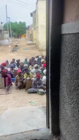 A group of approximately 30 children are sitting on the ground in a dusty street, facing a boy who is standing and holding a wooden board. The children are wearing traditional clothing, including colorful headscarves and robes. Some of the children have wooden boards in front of them, and they appear to be writing on them with sticks. The standing boy is wearing a yellow shirt and light-colored pants, and he is also holding a wooden board. The street is lined with buildings made of concrete and corrugated metal. Trees and utility poles are visible in the background. The sky is overcast, suggesting that it may be raining or about to rain. The overall scene depicts an outdoor classroom setting, likely in a developing country.