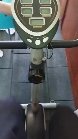 The video shows a person riding an exercise bike. The camera is positioned close to the handlebars, offering a first-person perspective. The bike has a display screen with several digital readouts. Other people are visible in the background, including one seated on a bench. The scene takes place in a gym with black rubber flooring. Weights and other exercise equipment are scattered throughout the room.