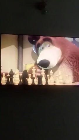 The video displays a television screen showing a scene from an animated show. A large brown bear is sitting at a table, playing chess. The bear is looking at the chessboard with a thoughtful expression. The bear picks up a chess piece and appears to be considering its next move. The background shows a cozy room with a window looking out onto a snowy landscape.
