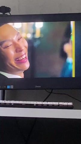 The video shows a close-up of a computer monitor displaying a scene from a movie or television show. In the foreground, a man with short dark hair and glasses is laughing and looking to his right. He is wearing a dark suit jacket over a white shirt. Behind him, slightly out of focus, another man with short dark hair and a wide grin is visible, also wearing a dark suit jacket. The background appears to be an indoor setting with lights and some indistinct figures. A webcam is mounted on top of the monitor. Below the monitor, a keyboard is visible on a white surface.