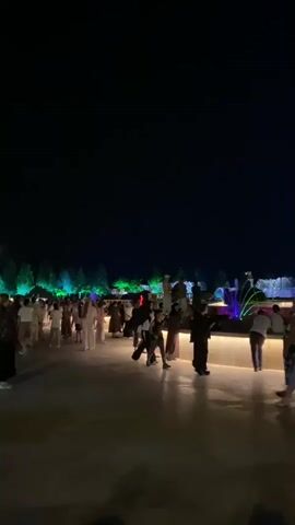 The video shows a nighttime outdoor scene with a dark sky overhead. In the foreground, a paved area is lit by artificial lights, reflecting on the ground. Several people are scattered across this area, some walking, some standing, and some sitting. They are dressed in casual and traditional attire. In the background, there are illuminated fountains with water jets spraying upwards, creating a dynamic display against the darkness. Lush green foliage and architectural structures are visible behind the fountains, also illuminated with colored lights, primarily green and blue. The overall atmosphere appears festive and social.