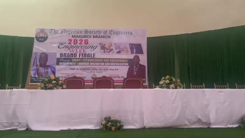 The media file shows a formal event setup, likely a conference or ceremony, in an indoor venue. The focal point is a long table draped in white cloth, with several chairs lined up behind it. In front of the table, a green, artificial turf covers the floor.

A large banner hangs prominently behind the table, bearing the text "The Nigerian Society of Engineers MAKURDI BRANCH 2026 Engineering WEEK GRAND FINALE". Below this, the theme is stated as "SMART TECHNOLOGIES FOR SUSTAINABLE DEVELOPMENT THROUGH INNOVATION AND DIGITALIZATION". The banner also features images of individuals, suggesting speakers or honorees. Two of these individuals are clearly visible in portraits on the banner.

To the left of the table, a podium with a projector is set up, indicating a presentation or speech is part of the program. To the right of the table, large black speakers and an air conditioning unit are visible. Floral arrangements, appearing to be yellow and white flowers, are placed strategically around the table and on the podium.

The backdrop consists of green draping that covers the walls and extends to the ceiling. Above the green drapes, a cream-colored ceiling is visible. The lighting suggests it is daytime or a well-lit indoor space, with no indication of weather conditions. The overall atmosphere appears professional and organized, fitting for an engineering society event. The presence of the banner and the formal seating arrangement point to a planned, significant occasion.