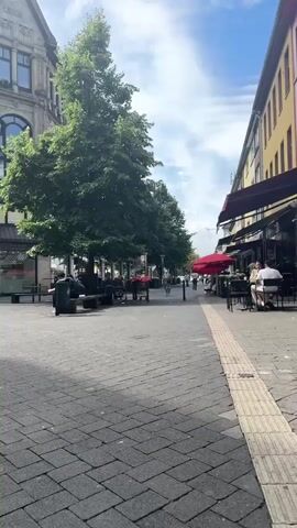 This video shows a pedestrian walkway in a European city on a sunny day with a few scattered clouds. The walkway is paved with interlocking bricks. On the left side of the walkway is a large tree, and behind it is a multi-story building with ornate architecture. Several people are sitting on benches under the tree. Further down the walkway, there are outdoor seating areas for cafes or restaurants, with red umbrellas providing shade. People are seated at tables. On the right side of the walkway, there are buildings with awnings and storefronts. In the distance, a few people are walking and cycling. A white delivery truck is parked on the far left side of the frame.