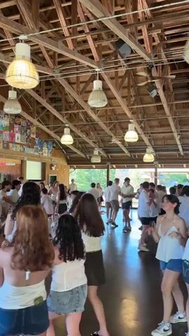 The video shows a group of young people dancing in a large, open room with a wooden ceiling and exposed beams. The room is lit by multiple pendant light fixtures. The walls are decorated with posters and what appear to be framed images. The floor is dark and reflective. Most of the people are wearing white tops and denim shorts. They are actively dancing and interacting with each other in a lively atmosphere. Some individuals are smiling and appear to be enjoying themselves. The overall scene suggests a casual gathering or party.