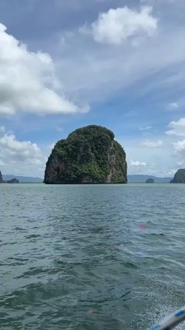 A drone shot pans right across a body of water with a large, green, rocky island in the center of the frame. The water is choppy and appears blue-green. The sky is blue with white, puffy clouds. The island is covered in lush green trees and vegetation, with steep, rocky cliffs visible on its sides. In the distance, to the right of the main island, are smaller, similarly vegetated islands. The shot is taken from the perspective of a boat, as indicated by a blurred rope or cable visible in the lower right corner of the frame. The drone moves slowly, revealing more of the surrounding islands and the expanse of the ocean.
