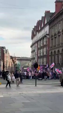 The scene captures a public gathering in an urban setting, with a crowd of people visible in the distance, many waving British flags. Several individuals are walking in the foreground across a paved area, while the background features traditional buildings. The sky is overcast.