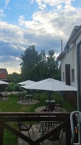 The video pans across an outdoor seating area with tables and umbrellas, surrounded by green grass and trees. A building with a chimney is visible in the background, as is a partially obscured red brick house with a peaked roof. The sky is blue with scattered clouds. There are no people visible in the video. The overall atmosphere appears to be calm and serene.