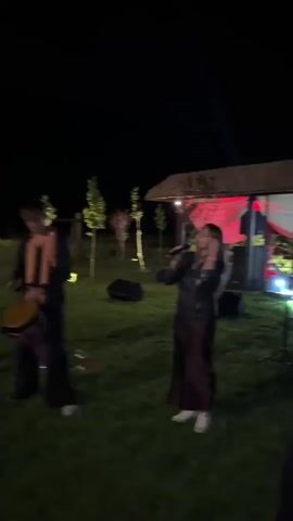 The video shows an outdoor nighttime performance. Four people are visible on a grassy area. On the left, a person wearing a dark jumpsuit is playing a flute. In the center, a young man in dark pants and a sleeveless shirt with an orange scarf is playing a hand drum. To the right of the drummer, a woman in a dark top and a long brown skirt is singing into a microphone. To the far right, a man with a beard is playing an accordion. Behind them, there is a wooden structure with red fabric draped inside, and trees are visible in the background. The ground is covered in grass.