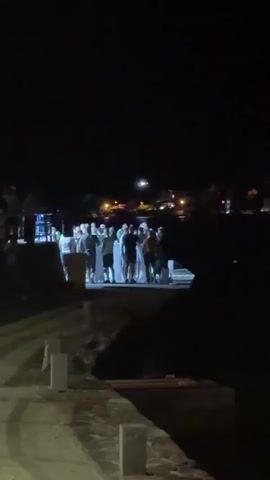 A group of people are standing on a raised platform at night. They appear to be facing a body of water, as there are lights and buildings visible across the water in the background. Some people are wearing white clothing. A railing is visible to the left of the group. The ground in the foreground is paved.