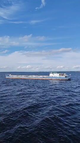 A cargo ship is sailing on a large body of water under a partly cloudy sky. The ship is long and narrow, with a white superstructure at the stern and a long, flat deck. The water is a deep blue with moderate waves. The sky is blue with scattered white clouds.