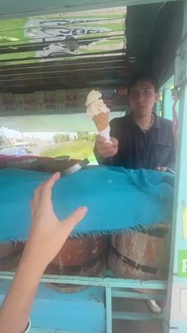 A person is buying ice cream from a vendor. The vendor, a young man with dark hair, is handing a triple-scoop ice cream cone to a customer across a teal-covered countertop. The ice cream appears to be a light color. The vendor is positioned inside a small, open-air stall with a turquoise and pink frame, showcasing flavor options on the wall above. The customer is identifiable by their arm reaching out to receive the cone, featuring a bracelet on their wrist. A second person is visible briefly outside the stall, partially obscured. The stall's base has wooden barrels used as a counter base. The surrounding environment appears to be an outdoor roadside setting, with hints of traffic and buildings in the background.