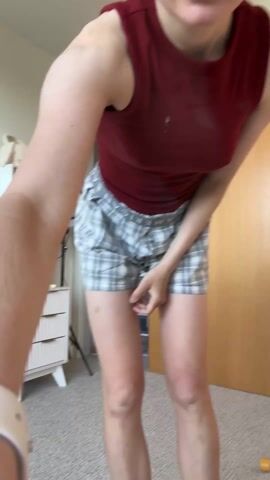 Here is a description of the video:
The video shows a woman in a bedroom. She is wearing a maroon sleeveless top and plaid boxer shorts. She is standing near a vanity with drawers and a ring light. Behind her is a closet with a wooden door. The woman puts her hands behind her head and stretches her back by slightly arching and bending to the side.
