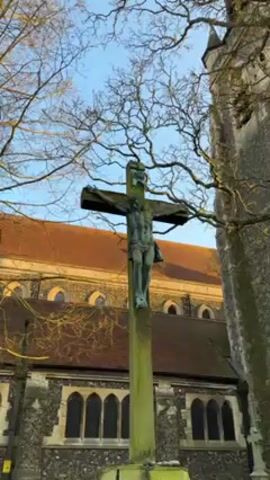 The video shows a crucifix with a statue of Jesus Christ affixed to it. The cross is made of wood and stands in front of a church building. The building is made of stone and has a tiled roof. There are trees surrounding the cross and church. The sky is light blue, and the sun is shining.