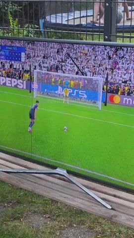 A person is recording a television screen outdoors. The screen shows a soccer game, specifically penalty kicks, with the score displayed as PSG and ARS tied at 0-0. A player in a dark jersey is taking a penalty kick. The game footage transitions to a shot of José Mourinho, a soccer coach, on the sidelines. The television is set on a wooden bench outside, near some grass and a fence. A trampoline is visible behind the fence with people on it.