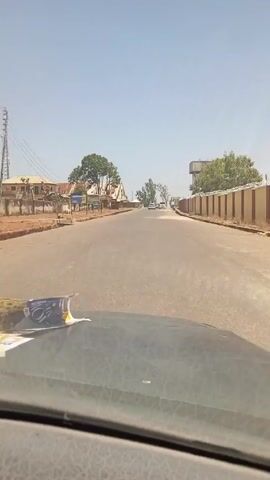 The video shows a view from inside a vehicle driving down a road on a bright, clear day. The road is paved and appears to be relatively empty, with a few cars visible in the distance. On either side of the road, there are buildings and a wall topped with barbed wire. A water tower can be seen in the distance to the right of the road. The vehicle's dashboard is visible in the foreground. There is a folded newspaper or magazine on the dashboard. The overall scene suggests a quiet, possibly rural, setting.