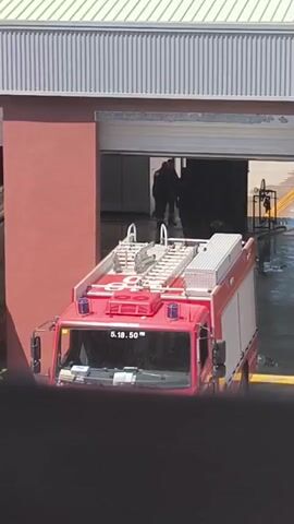 The video captures an exterior view of what appears to be a fire station, with two red and white fire trucks. In the foreground and right, a fire truck is partially visible, parked either in or near a bay. The truck features "5.18.50" on its windshield and the word "bombers" (Catalan for firefighters) on its front bumper. Blue emergency lights are visible on its roof.

To the left, another red and white fire truck is mostly obscured by a red wall, with only a portion of its side and top visible. The building itself has prominent red walls and a gray, corrugated metal roof. Inside the bay, where the right-hand truck is positioned, two individuals are discernible in the background, largely silhouetted. There is visible water on the ground within the bay, suggesting that a cleaning or maintenance activity, such as washing the trucks, may be taking place. The scene is brightly lit, indicating daytime. The context suggests the location is likely in Vilafranca del Penedès, Spain.