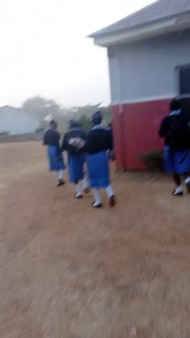 A group of students in blue skirts and dark tops walk on a dirt path away from the camera. A building with a red and grey exterior is on the right. The students are moving away from a grassy area with trees in the distance, under a hazy sky. Some students are carrying books or notebooks. At least one student has a backpack. The ground is mostly dirt with some small patches of grass.