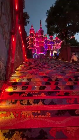 The video shows a colorful mosaic staircase leading up to a castle-like structure lit with multicolored lights.  The steps are decorated with shards of red, yellow, black, and gray tiles. Red neon lights line a brick wall to the left of the staircase.  Small lights illuminate the edges of the steps. At the top of the stairs, the castle glows with red, pink, blue, green, and yellow lights. People can be seen walking around the base of the structure.  A large tree is visible on the right side of the frame. The sky is dusky.