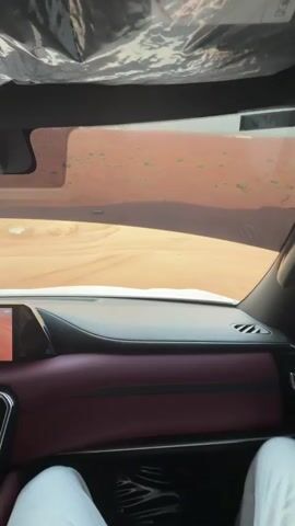 From inside a vehicle, a video shows driving through a sandy desert. The dashboard is maroon. There is a person in white clothes in the passenger seat. A windshield allows a view of the sand dunes. The camera bobs slightly as the vehicle bounces. There are a few green shrubs spotted in the sand. The sky is light blue.