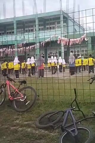 A group of people, likely students, are assembled on a grassy field in front of a green building under construction. They are mostly wearing yellow vests over dark clothing, and some are wearing hijabs. The building has multiple floors and visible scaffolding. The field is enclosed by a chain-link fence. Several bicycles are visible in the foreground, some leaning against the fence and others lying on the ground. Red and white decorative banners hang across the front of the building. The sky is overcast.