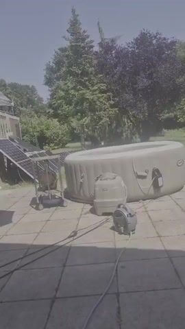 The video shows an outdoor scene with a large inflatable hot tub filled with water. Next to the hot tub are a solar panel array tilted towards the sun and what appears to be a water pump connected to the hot tub by hoses. The ground is paved with square tiles, and trees and foliage are visible in the background under a bright sky. The hot tub is emitting a faint mist, suggesting it may be heated.