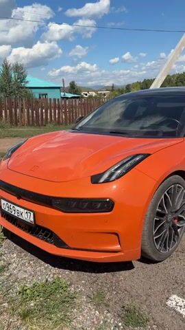 The video begins by showing the front quarter of an orange electric car parked outdoors. The car has a black roof and black rims. The scene then pans to the right, revealing the side of the car and an electric charging station. The charging cable is plugged into the car. The charging station is labeled "РОССЕТИ ЭЛЕКТРОСЕТИ". The background consists of a blue sky with white clouds, trees, a wooden fence, and a building. The camera continues to pan right, focusing on the charging process. Finally, the video zooms in on the driver's side door handle as a hand reaches for it.