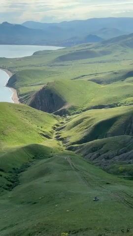 The video depicts a landscape featuring rolling green hills descending towards a coastline. In the distance, there's a body of water, presumably a sea or a large lake, with a city or town visible along its edge, nestled between the water and distant mountain ranges. The sky is partly cloudy, with the sun breaking through in sections. The foreground consists of grassy hills, with a dirt path winding down a prominent valley. Small figures of people are seen walking along the path. A motorcycle is stationary at the bottom of one of the hills, near the path.