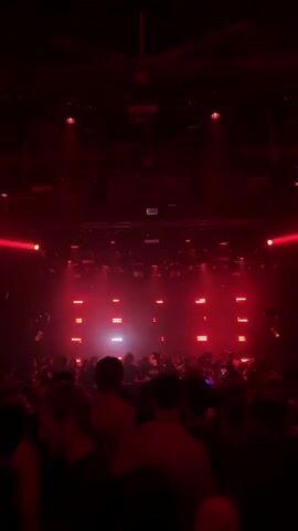 The video shows a crowded indoor venue, likely a club or concert hall, bathed in red and pink lighting. A large crowd of people is visible from behind, facing a stage or performance area. The lighting is dynamic, with beams of red light cutting through the darkness and rectangular light panels illuminating the stage in a rhythmic pattern. Disco balls are also visible hanging from the ceiling, reflecting the light. The atmosphere appears energetic and festive, with people gathered to watch a performance. The focus is on the visual spectacle of the lights and the collective experience of the crowd.