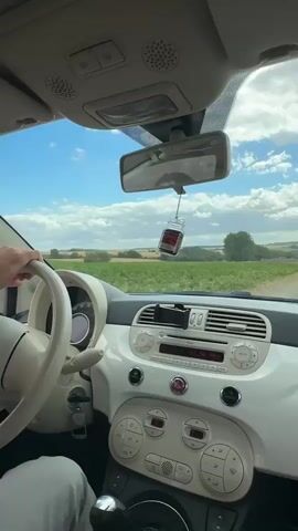 The video is shot from inside a car, showing the driver's hands on the steering wheel and the dashboard. The driver is wearing a white shirt. Outside the car, there is a field of green crops and a blue sky with clouds. A car air freshener, shaped like a small glass bottle with a red liquid, is hanging from the rearview mirror. The dashboard is white and cream colored.