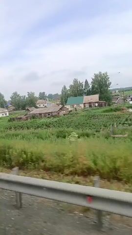 The video shows a rural landscape with several small houses scattered across a green, grassy hill. The houses appear to be made of wood and have simple structures, some with metal roofs. There are trees and bushes interspersed among the houses and on the surrounding terrain. The sky is cloudy, suggesting an overcast day. In the foreground, a gray metal barrier or railing is visible, indicating that the footage was likely captured from a moving vehicle traveling along a road. The overall impression is of a quiet, rustic village scene.