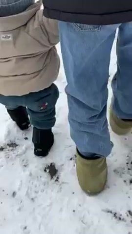 A toddler in a beige jacket, teal pants, and black boots walks with an adult through the snow. The adult wears jeans and green boots. The toddler holds the adult’s hand, and they walk slowly across the snowy ground.