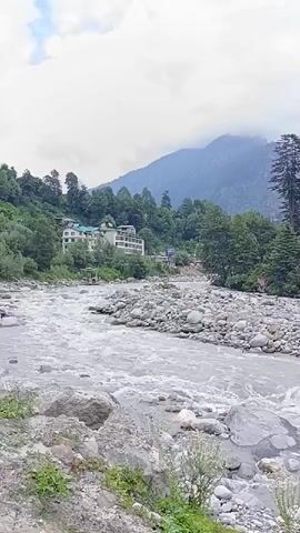 This is a vertical, low-angle video of a fast-flowing river with a rocky bed. On the left bank, there is a multi-story hotel with a green roof, surrounded by trees. The river continues past the hotel and towards a mountain range covered in lush green trees. The sky is overcast with clouds, and the overall impression is one of a scenic, natural landscape. There are no people visible in the video.