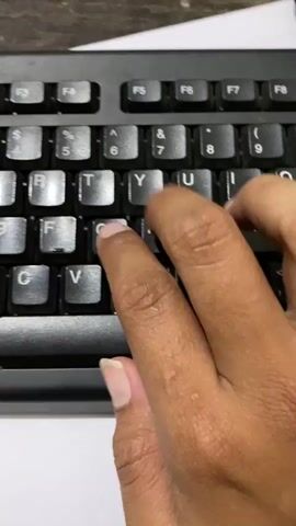 The video shows a close-up of a person's hands typing on a black computer keyboard. The keyboard has standard QWERTY layout keys and function keys at the top. The person's right hand is primarily visible, with fingers pressing down on keys such as 'R', 'T', 'F', and 'C'. The background is blurred but appears to be a desk with a white surface and a dark textured surface, possibly wood or laminate. The lighting is bright, highlighting the keyboard and hands. The video is short and focuses on the action of typing.